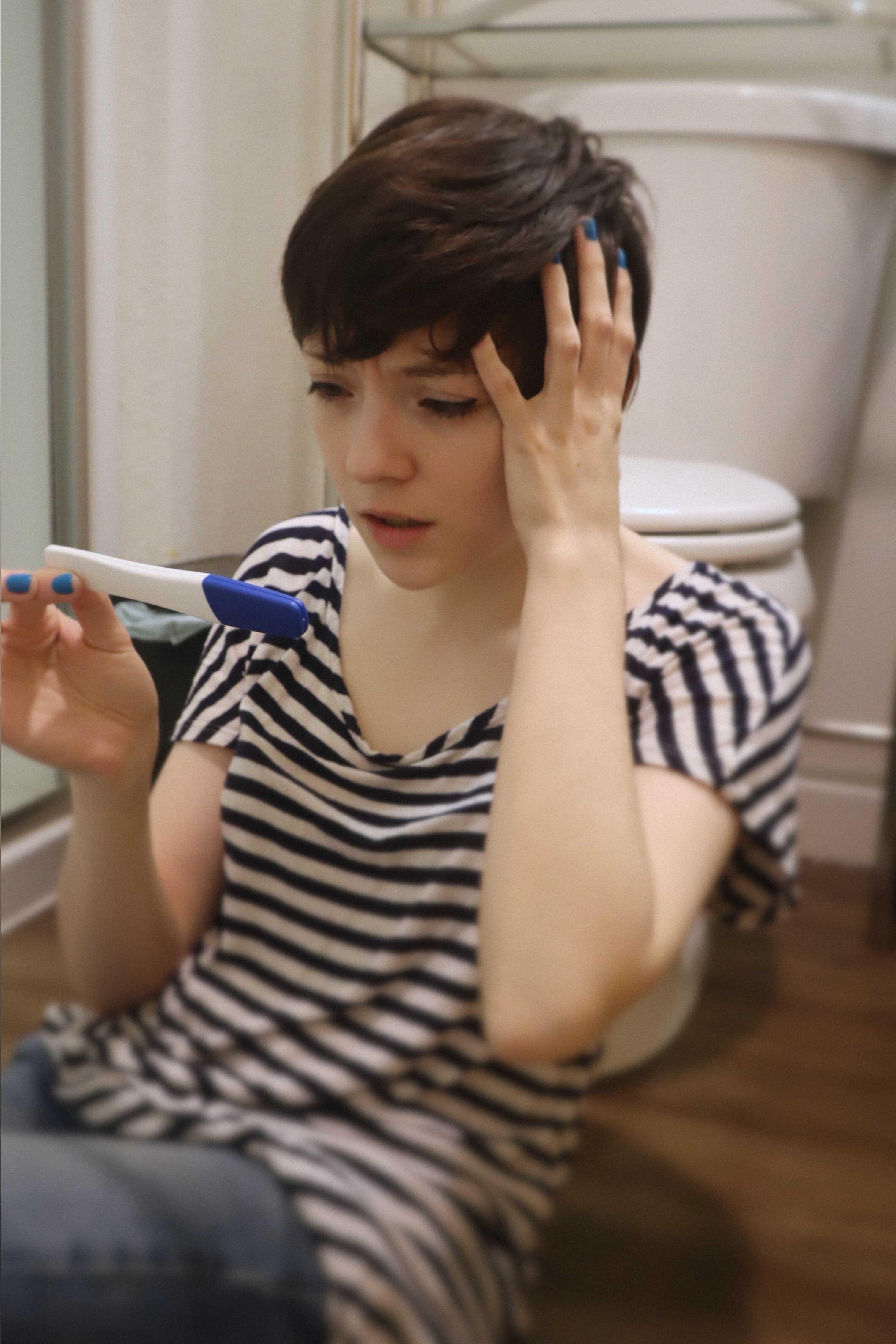 Eine weiblich gelesene Person sitzt vor einer Toilette auf dem Fußboden und hält einen Schwangerschaftstest in der einen Hand während sie sich mit der anderen in die Haare greift und dabei mit leicht geöffnetem Mund nicht besonders erfreut auf den Test schaut